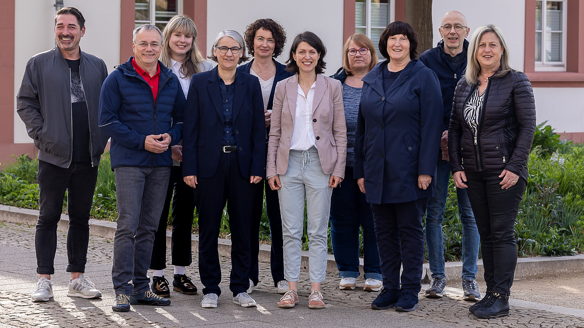 Eine Gruppe von zehn Erwachsenen, fünf Männer und fünf Frauen, steht im Freien vor einem Gebäude, posiert für ein Gruppenfoto und lächelt in die Kamera.