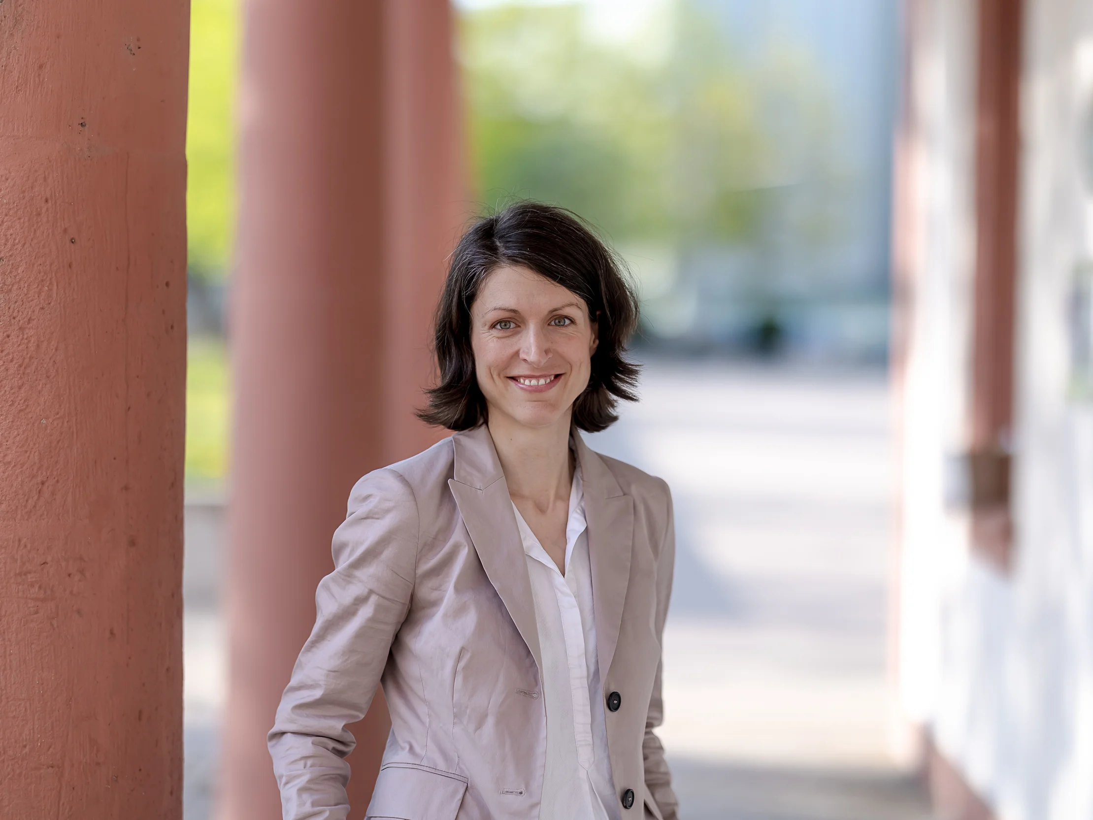 Eine Frau mit kurzen braunen Haaren, die einen hellbeigen Blazer und ein weißes Hemd trägt, steht im Freien und lächelt neben einer roten Säule. Der Hintergrund ist unscharf.