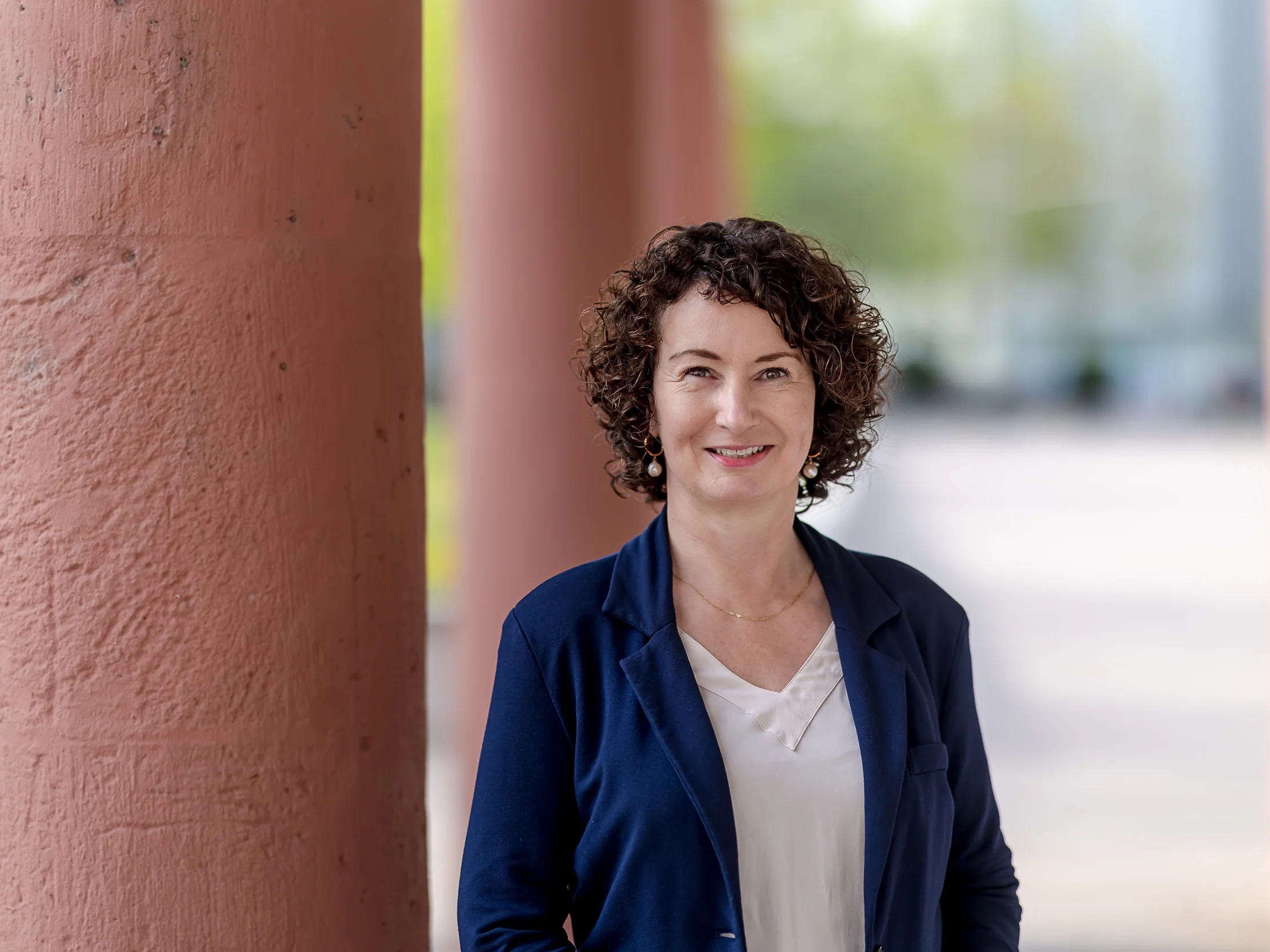 Eine Frau mit lockigem braunem Haar, die einen marineblauen Blazer und ein weißes Hemd trägt, steht im Freien neben einer großen rötlichen Säule und lächelt in die Kamera.