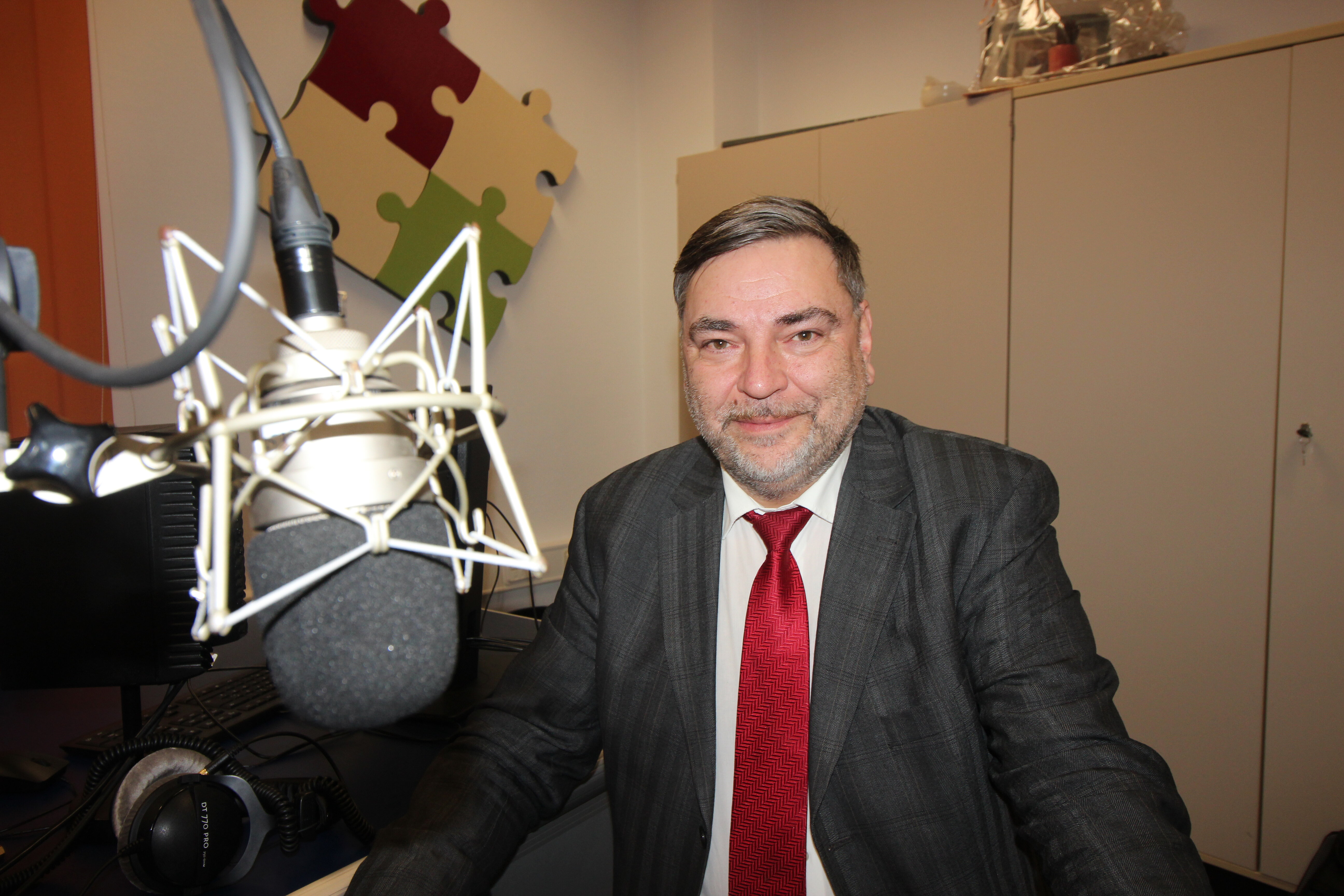 Ein Mann im Anzug und mit roter Krawatte sitzt an einem Schreibtisch vor einem großen Mikrofon in einem Aufnahmestudio mit Schränken und Puzzlestück-Wanddekor im Hintergrund.