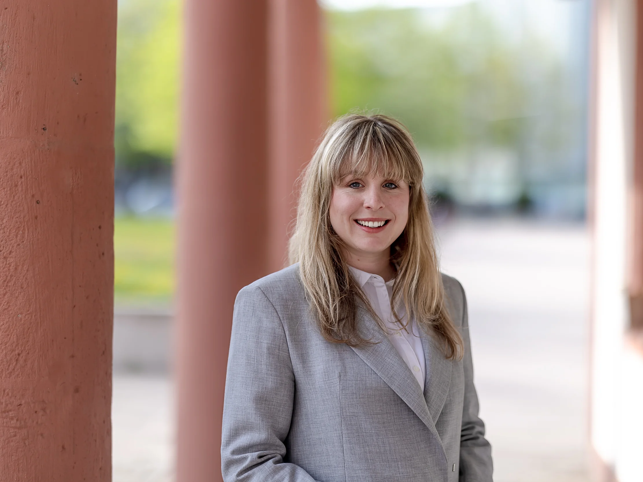 Eine Frau mit langen blonden Haaren und einem hellgrauen Blazer steht draußen neben großen Säulen und lächelt in die Kamera.