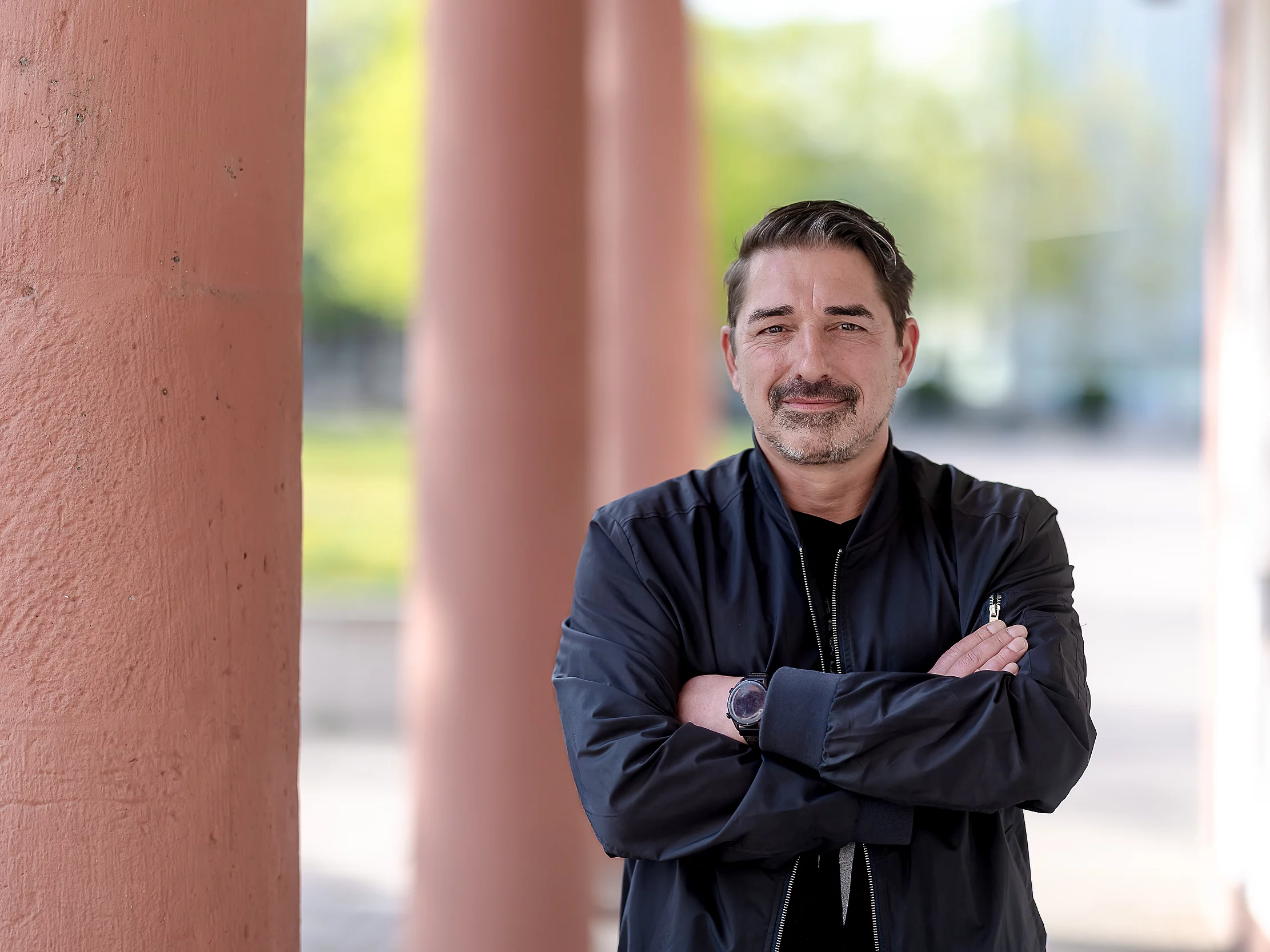 Ein Mann mit dunklem Haar und gestutztem Bart steht draußen mit verschränkten Armen und schwarzer Jacke vor rosa Säulen und einem unscharfen grünen Hintergrund.