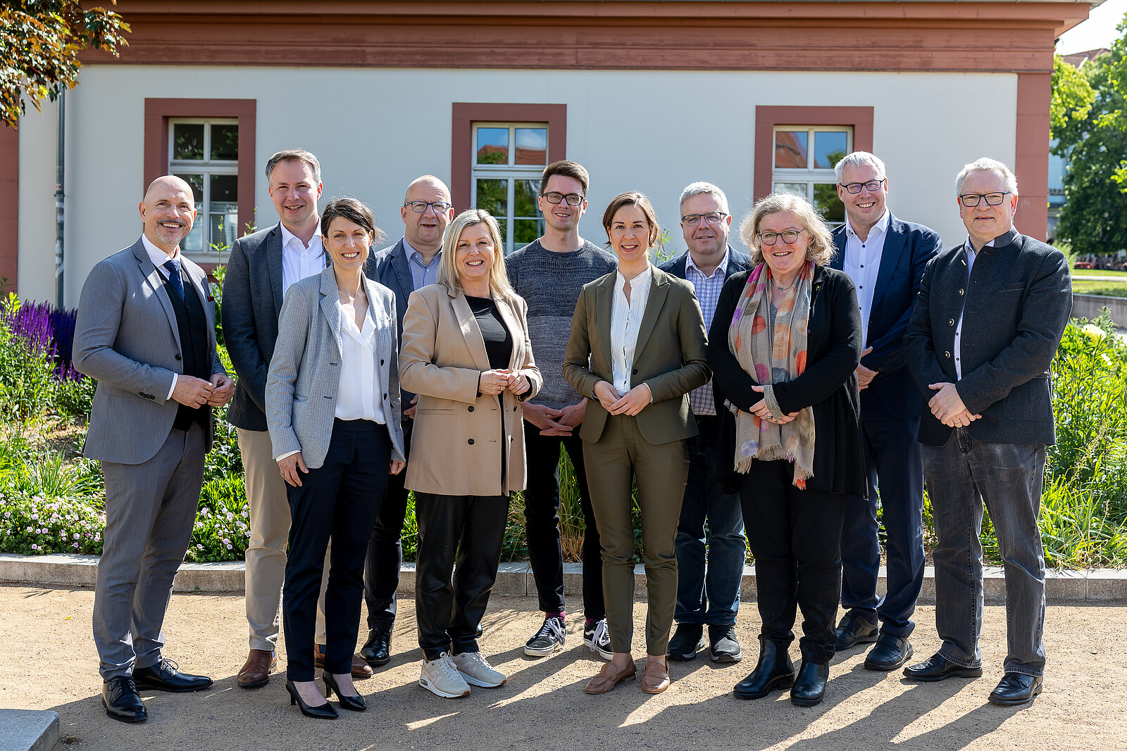 Eine Gruppe von elf Erwachsenen posiert und lächelt an einem sonnigen Tag im Freien vor einem Gebäude mit weißen Wänden und roter Verkleidung zusammen.