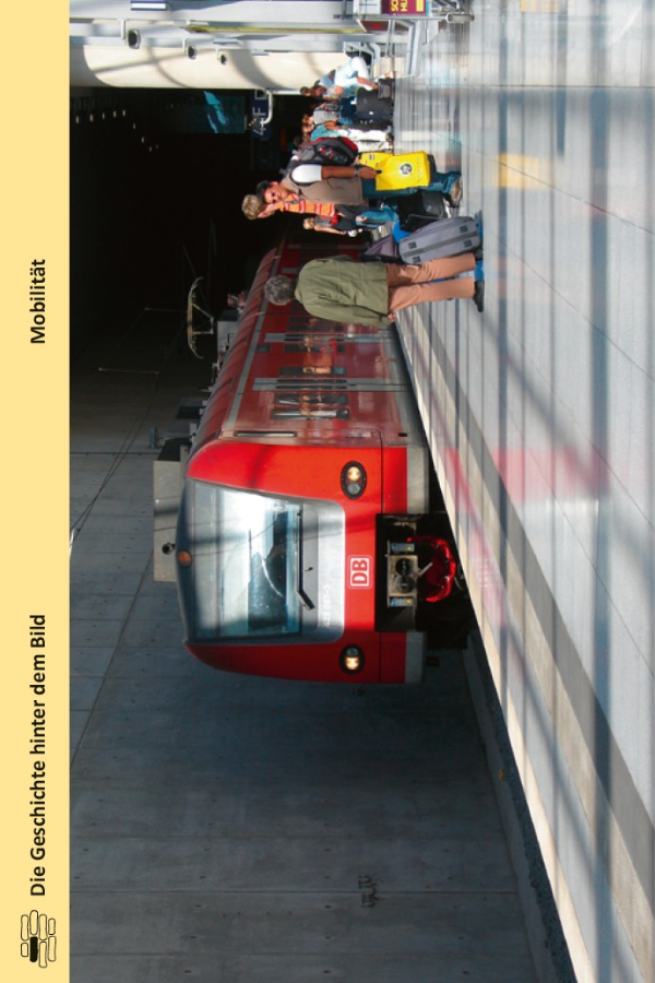 Ein roter DB-Zug hält an einem überdachten Bahnsteig. Mehrere Fahrgäste mit Gepäck gehen oder stehen in der Nähe. Der Bahnsteig ist sauber und gut beleuchtet, und die Szene lässt auf Reisen oder Pendeln schließen.