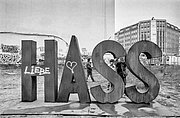 In großen Metallbuchstaben steht "HASS" mit Graffiti "LIEBE" und einem Herz auf dem ersten Buchstaben; zwei Personen stehen zwischen den Buchstaben in einer städtischen Umgebung mit Gebäuden und Graffiti.