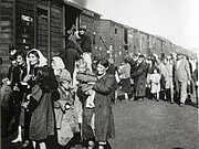 Eine Gruppe von Menschen, darunter Frauen und Kinder, geht neben einer Reihe von Eisenbahnwaggons entlang, während im Hintergrund Rauch aufsteigt.