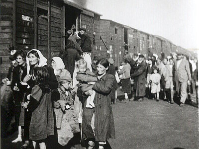 Eine Gruppe von Menschen, darunter Frauen und Kinder, geht neben einer Reihe von Eisenbahnwaggons entlang, während im Hintergrund Rauch aufsteigt.