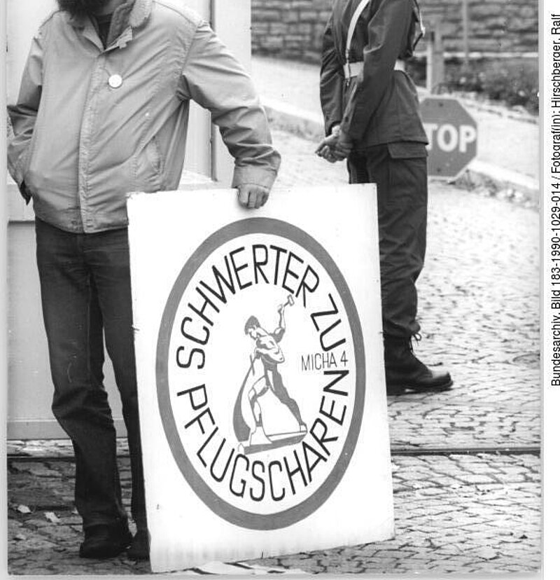 Ein bärtiger Mann steht mit einem Schild mit der Aufschrift "Schwerter zu Pflugscharen" neben einem uniformierten Wachmann auf einer Kopfsteinpflasterstraße.