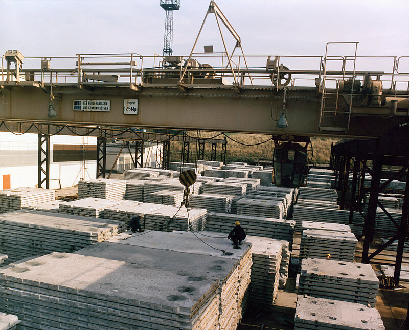 Ein Kran bewegt eine große Betonplatte über Stapel ähnlicher Platten in einem Industrielager, wobei zwei Arbeiter mit Schutzhelmen auf dem Boden zu sehen sind.