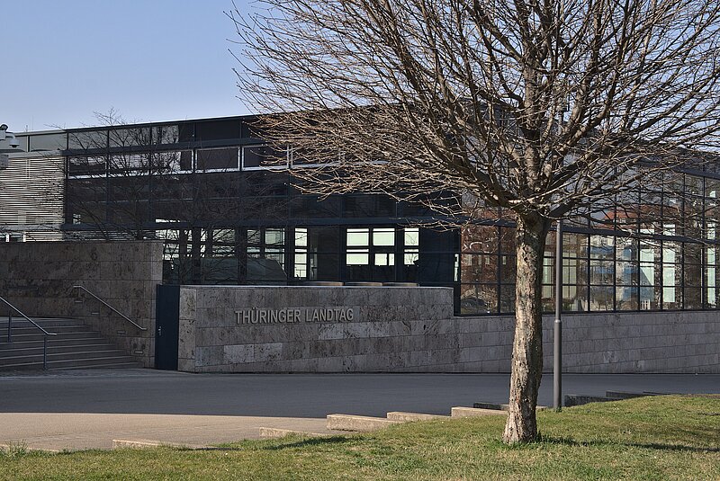 Ein modernes Gebäude aus Stein und Glas mit dem Schild "Thüringer Landtag" an der Wand, teilweise verdeckt durch einen blattlosen Baum im Vordergrund.