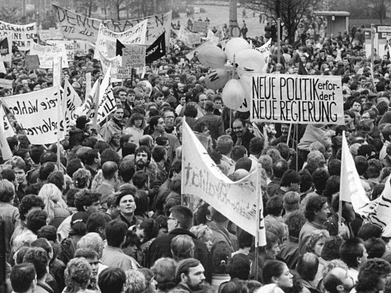 Fotografie einer großen Menschenmenge während der Friedlichen Revolution. Die Demonstrierenden versammeln sich im Freien und halten Schilder und Transparente mit Forderungen nach politischem Wandel und einer neuen Regierung.