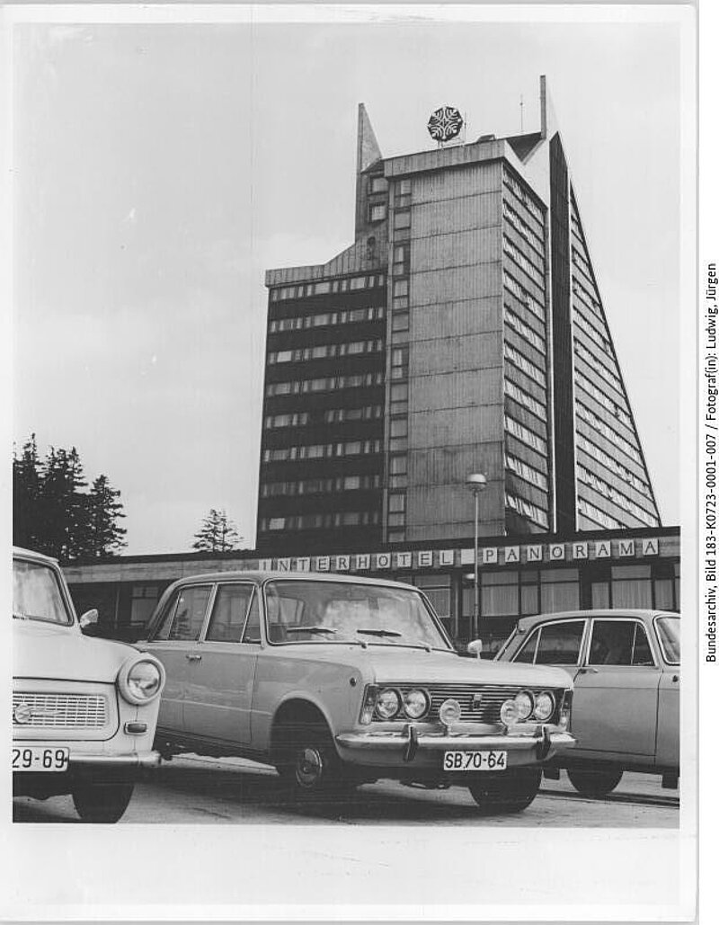 Drei Autos parken vor dem Interhotel Panorama, einem hohen, modernistischen Gebäude mit kantigem Design und einem Wappen auf der Spitze.