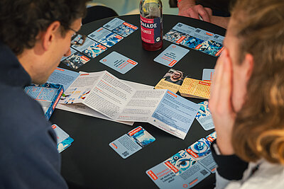 Zwei Personen sitzen an einem Tisch und lesen eine Broschüre, umgeben von verschiedenen Spielkarten und einer Flasche Limonade.