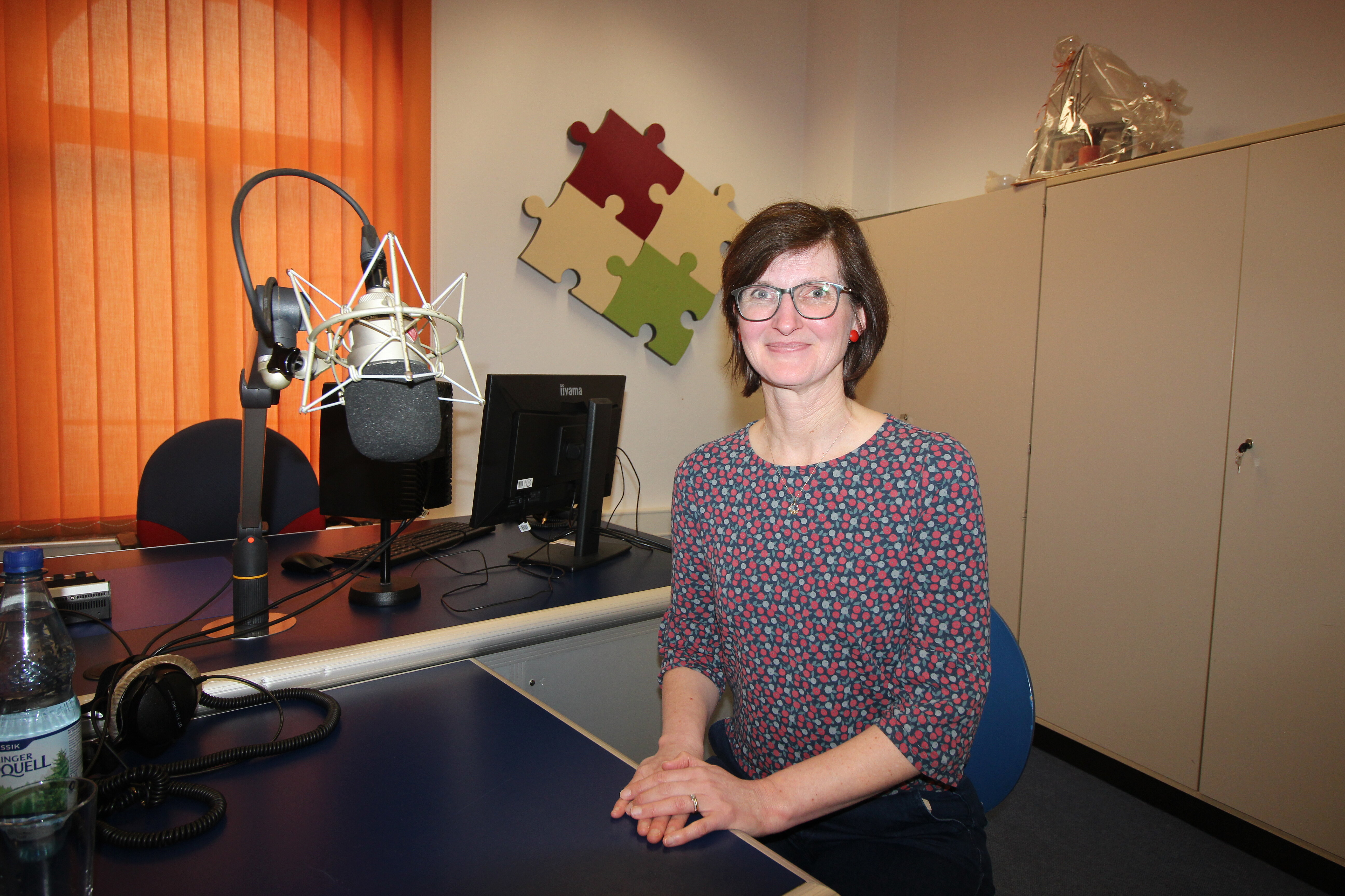 Eine Frau sitzt an einem Schreibtisch in einem Aufnahmestudio mit einem Mikrofon, einem Computermonitor und Kopfhörern in der Nähe. Eine Wasserflasche und ein Puzzlestück an der Wand sind zu sehen.