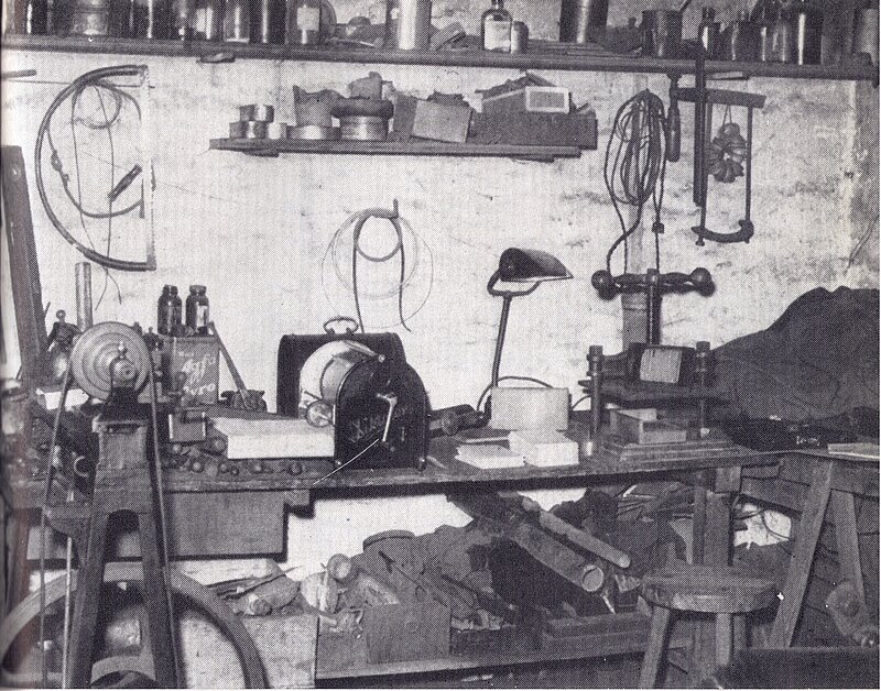 Eine überfüllte Werkstatt mit verschiedenen Werkzeugen, Maschinen, Regalen mit Gläsern und Dosen, einer Werkbank, einem Hocker und verstreuten Materialien.