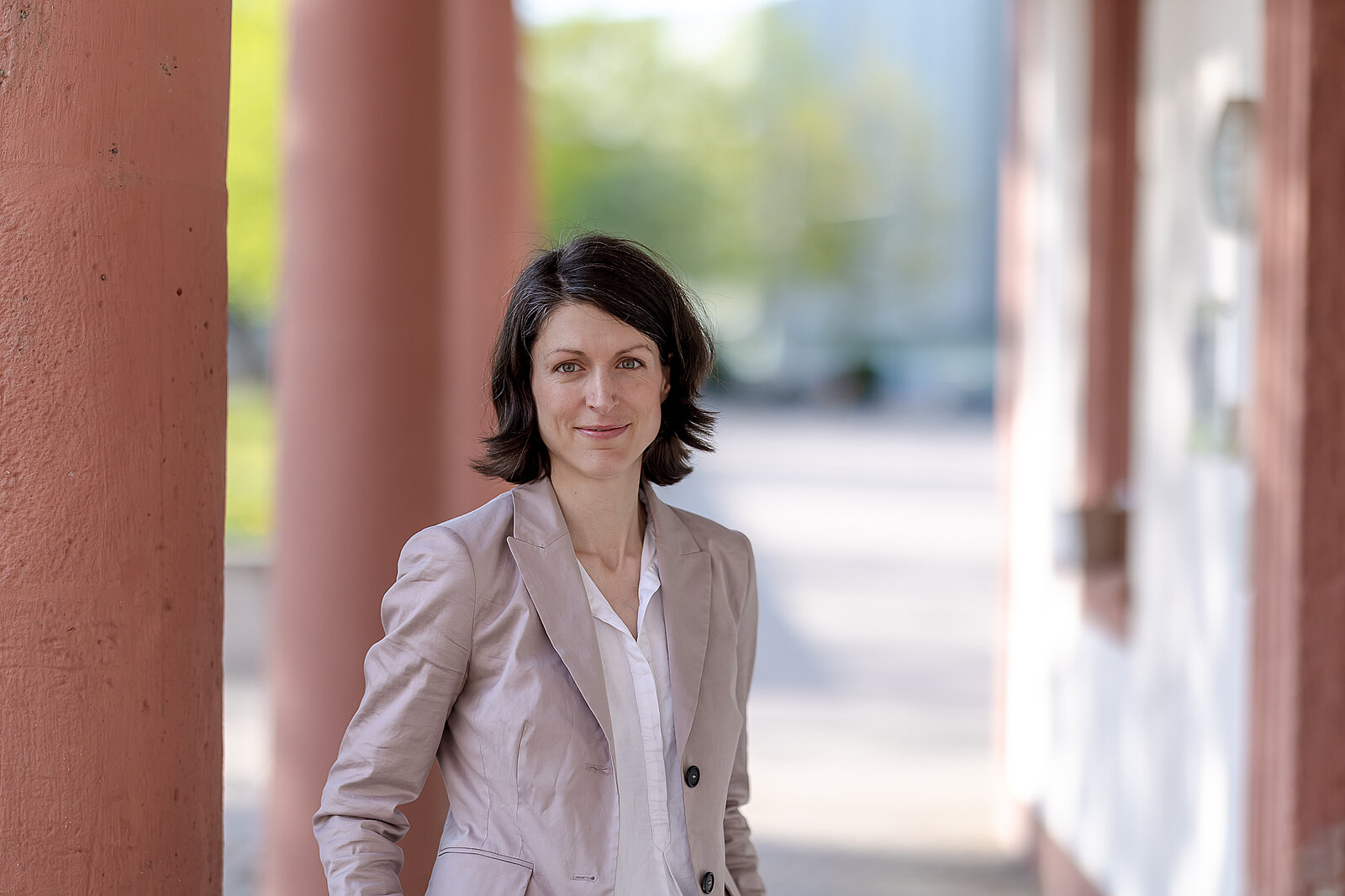 Eine Frau mit dunklem Haar, hellem Blazer und weißem Hemd steht im Freien in der Nähe roter Säulen und schaut in die Kamera.