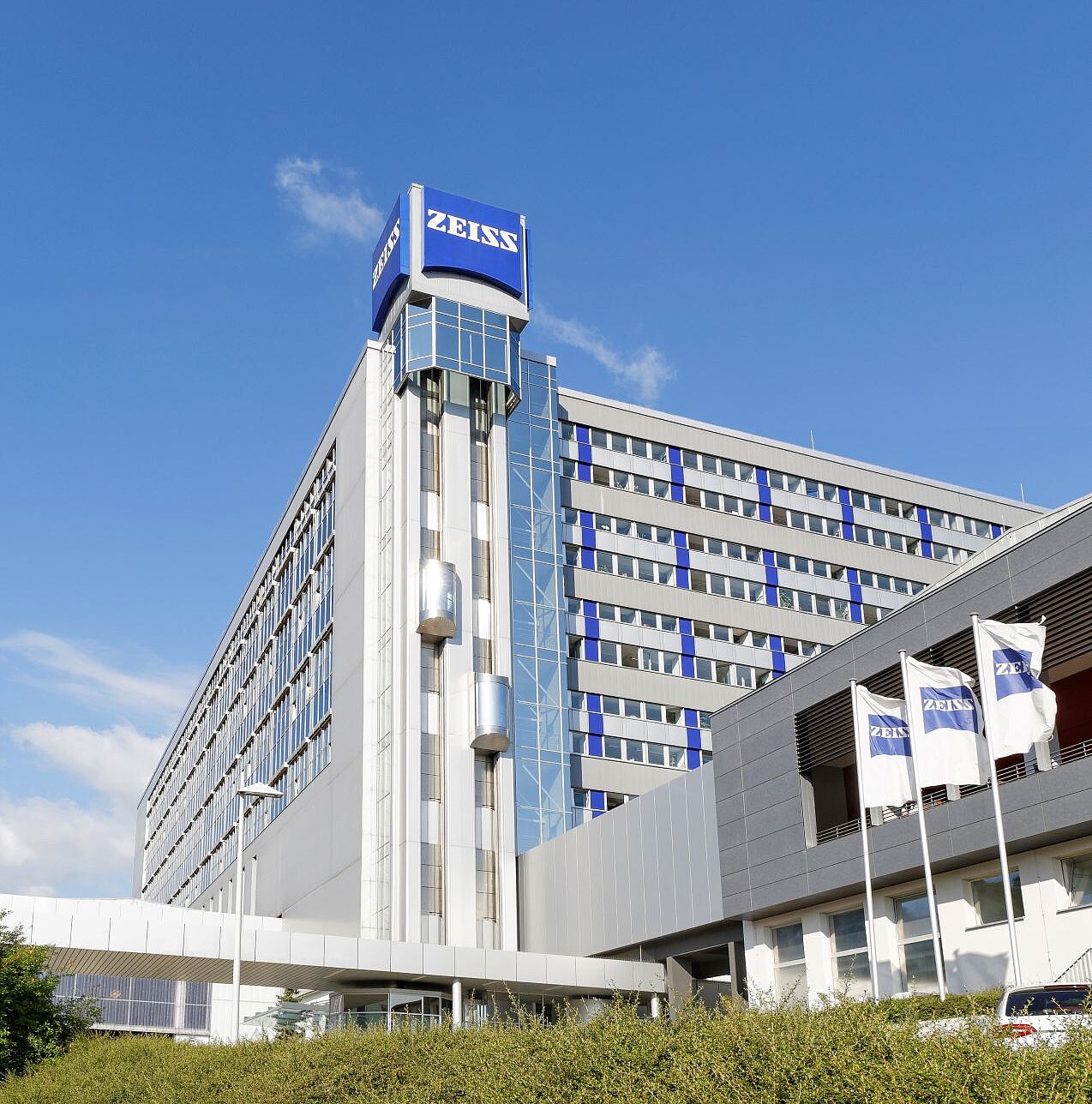 Modernes mehrstöckiges Bürogebäude mit "ZEISS"-Schriftzug, blauem Himmel darüber und drei weißen Fahnen davor, teilweise verdeckt durch Büsche.