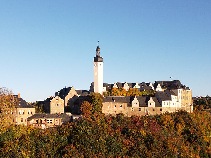 Ein großes historisches Schloss mit einem hohen Uhrenturm steht auf einem Hügel, umgeben von Bäumen, die sich unter einem klaren blauen Himmel herbstlich verfärben.