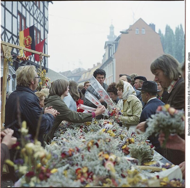 Menschen versammeln sich an einem Marktstand, an dem Sträuße und Blumenarrangements angeboten werden; im Hintergrund ist die ostdeutsche Flagge zu sehen.