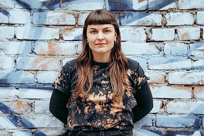 Eine Frau mit langen braunen Haaren und Pony steht vor einer bemalten Ziegelwand, trägt ein schwarzes T-Shirt mit Bleichmittelmuster und eine schwarze Hose und schaut in die Kamera.