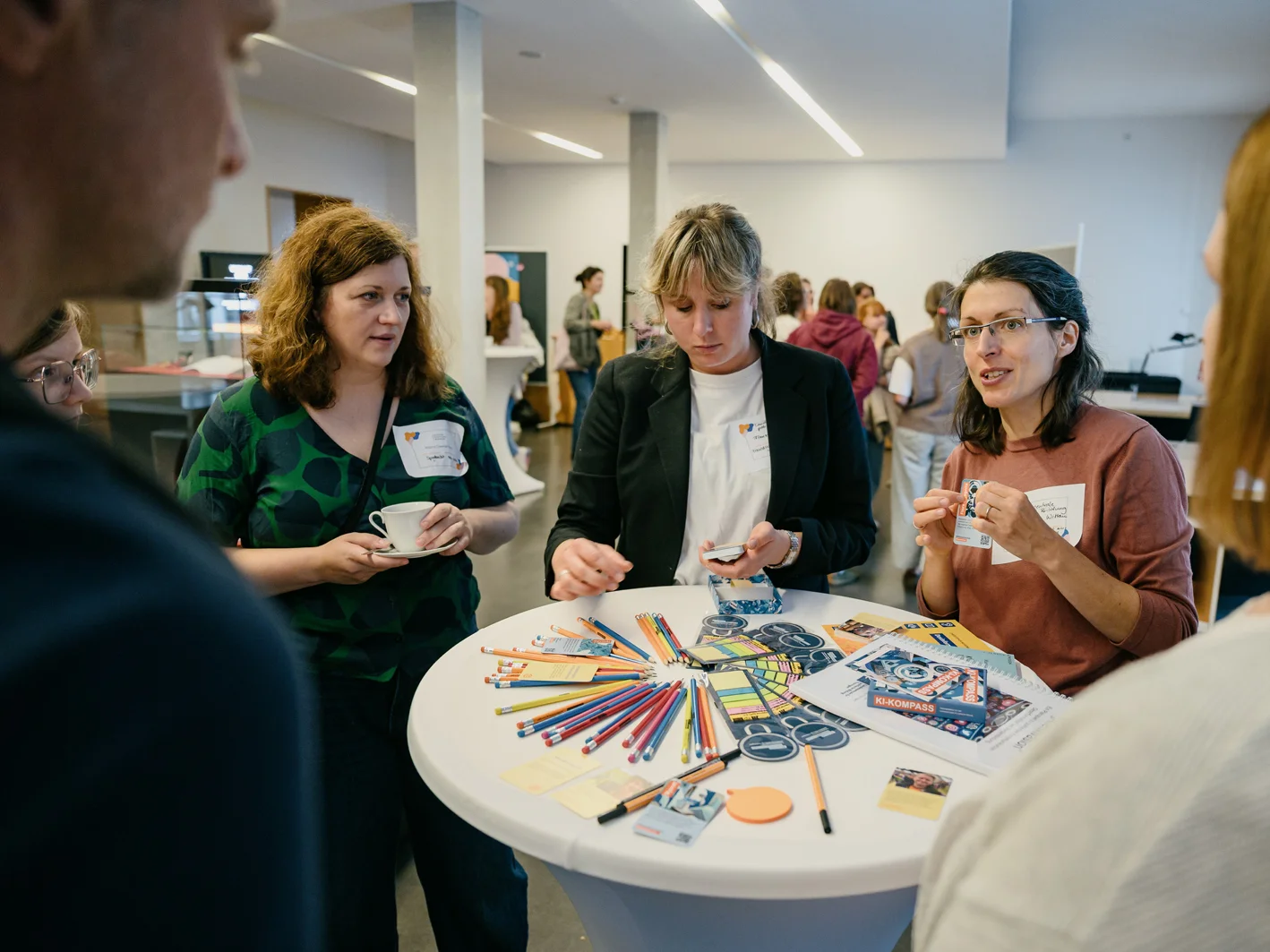 Vier Personen stehen in einem hell erleuchteten Innenraum um einen Tisch herum, der mit Malutensilien, Bleistiften, Scheren und Papieren bedeckt ist, und führen eine Diskussion.