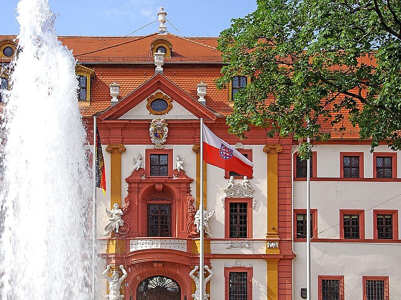Ein großer Brunnen vor einem historischen Gebäude mit rotem Dach, verzierter Fassade und mehreren Fahnen, darunter eine mit einem Wappen.