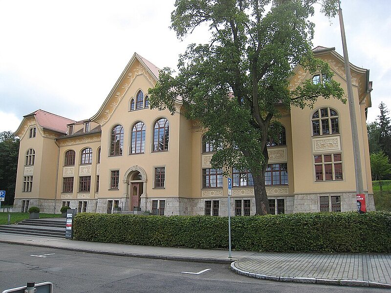 Dreistöckiges, beiges historisches Gebäude mit Rundbogenfenstern und rotem Dach, umgeben von Bäumen und einer Hecke entlang eines Bürgersteigs.