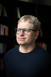Ein Foto des Autors des Buches, Stefan Wellgraf. Ein Mann mit kurzen blonden Haaren und Brille, der einen dunklen Pullover trägt, steht in einem Raum vor einem Bücherregal mit unscharf abgebildeten Büchern.
