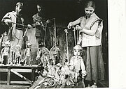 Drei Puppenspieler manipulieren Marionetten auf einer Bühne, wobei jeder von ihnen Fäden in der Hand hält, um detaillierte, menschenähnliche Puppenfiguren in einer Theatervorstellung zu steuern.