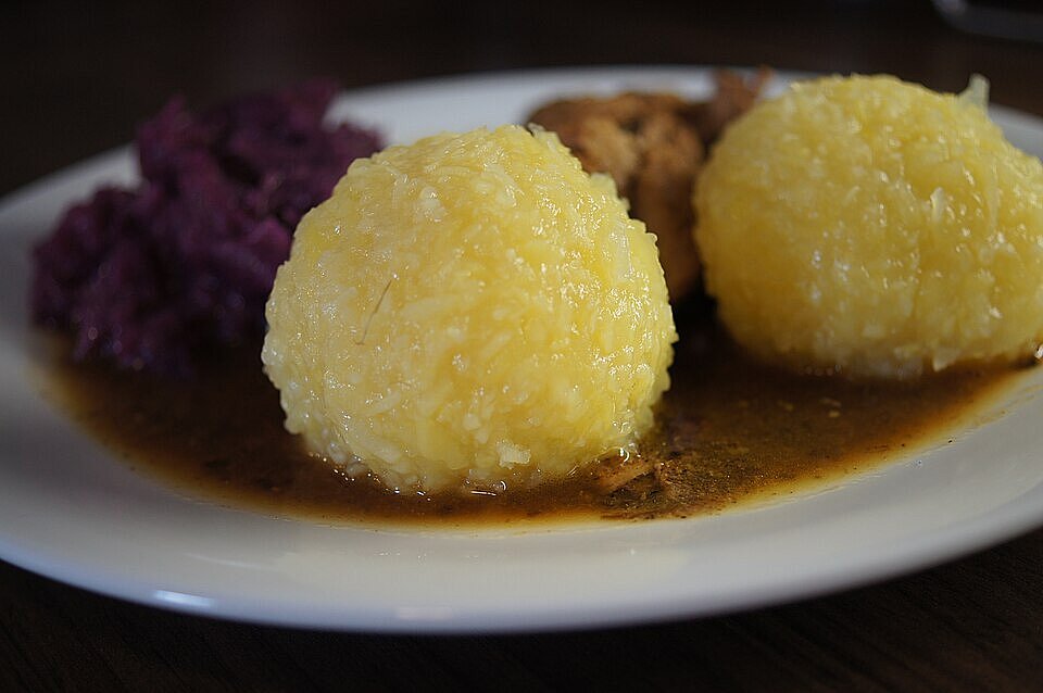 Thüringer Klöße mit Rindergulasch, Bratensoße und Rotkohl. Drei große, gelbliche Kartoffelklöße liegen auf einem Teller in dunkler Soße; dahinter sind Rindergulasch und Rotkohl zu sehen.