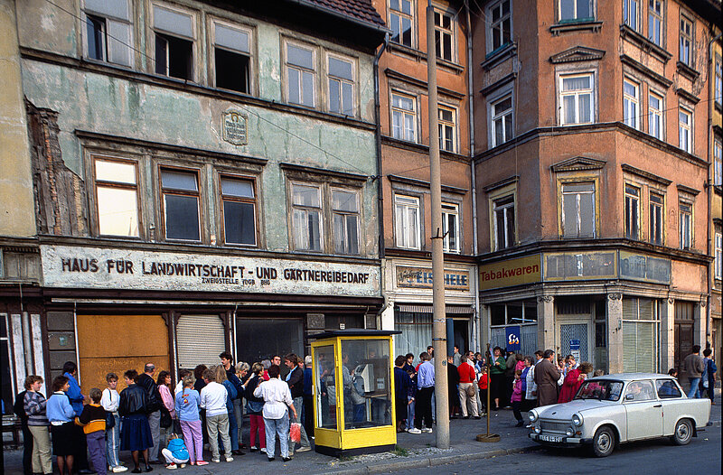 Eine Menschenmenge steht in einer langen Schlange auf einem Gehweg in der Nähe alter Gebäude, einer gelben Telefonzelle und eines geparkten weißen Autos.