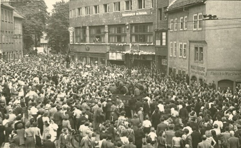 Drei Schwarz-Weiß-Fotografien zeigen: verstreute Papiere und Menschen auf einer Straße, eine Straßenbahn, die von Rauch umgeben ist, und eine große Menschenmenge in einem städtischen Gebiet, alle montiert auf einem linierten Archivbogen.