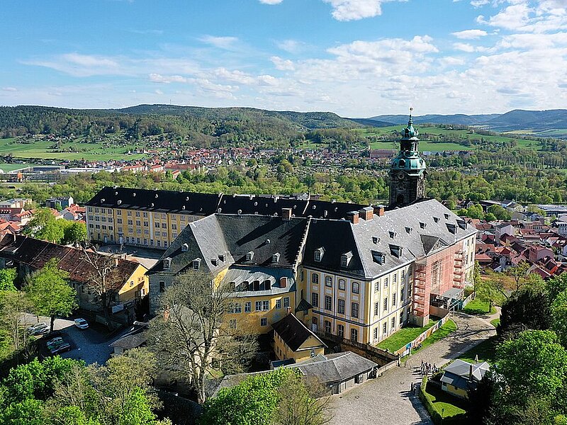 Luftaufnahme eines großen gelben barocken Gebäudekomplexes mit einem hohen Uhrenturm, umgeben von Grünflächen und einer kleinen Stadt, mit Hügeln im Hintergrund.