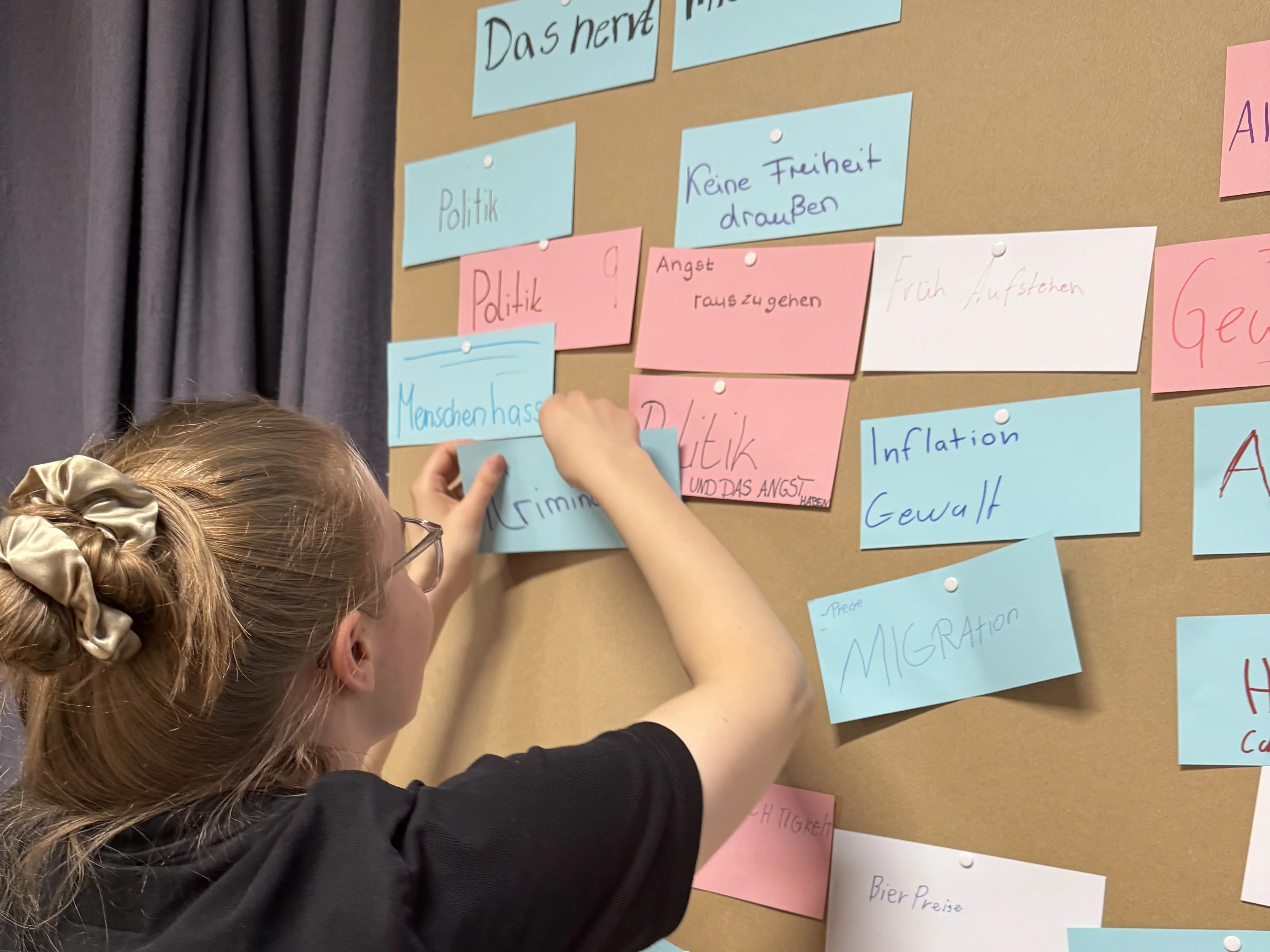 Eine Person ordnet handgeschriebene Karten mit deutschem Text auf einer braunen Wand an und ordnet die Antworten unter einer Überschrift, die lautet "Das nervt mich aktuell am meisten".