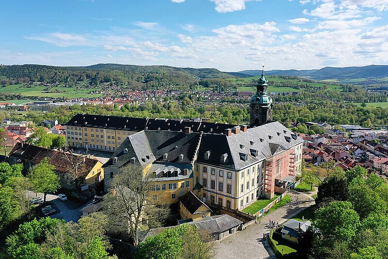 Luftaufnahme eines großen gelben barocken Gebäudekomplexes mit einem hohen Uhrenturm, umgeben von Grünflächen und einer kleinen Stadt, mit Hügeln im Hintergrund.