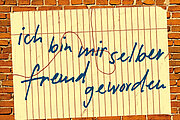 Ein handgeschriebener Zettel in deutscher Sprache auf liniertem Papier mit der Aufschrift "Ich bin mir selbst freundlich geworden" klebt an einer Backsteinmauer.