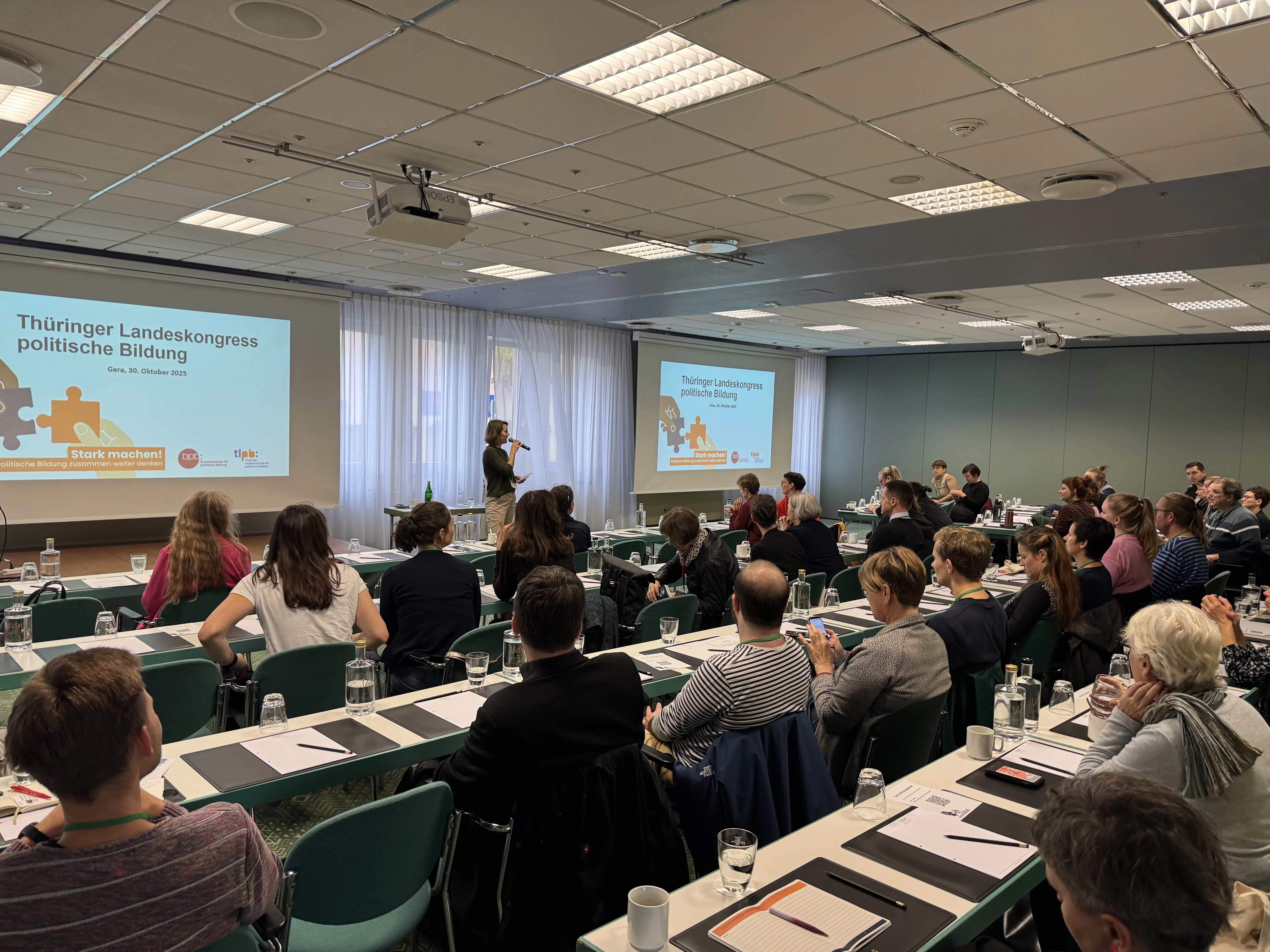 Ein Tagungsraum mit Teilnehmern, die an Tischen sitzen und einem Referenten gegenübersitzen, der auf einer Leinwand Folien zur politischen Bildung beim Thüringer Landeskongress präsentiert.