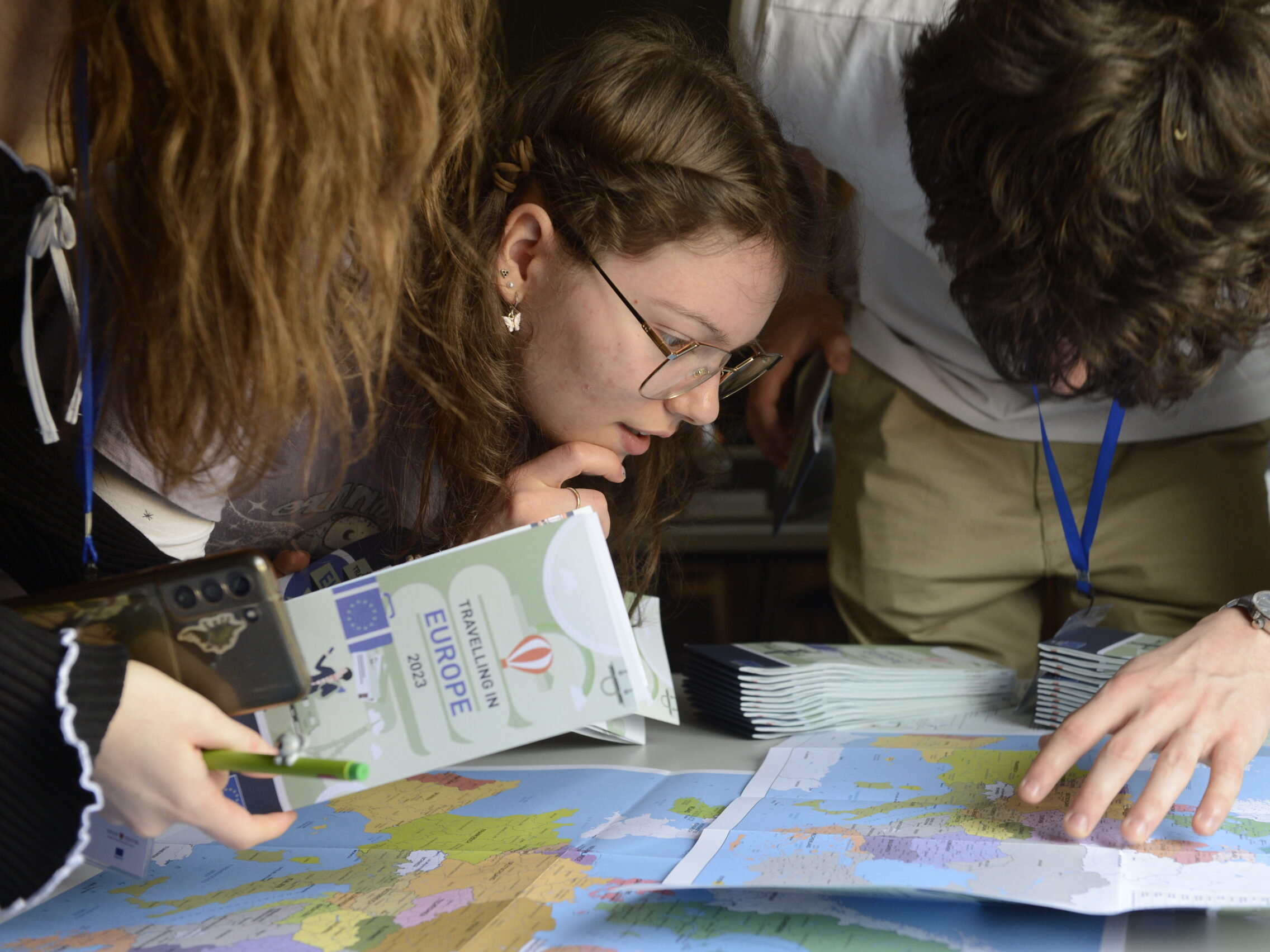 Drei Personen sehen sich eine Europakarte genau an, wobei eine einen Reiseführer mit dem Titel "Europa" in der Hand hält und die anderen auf Orte auf der Karte zeigen oder diese betrachten.