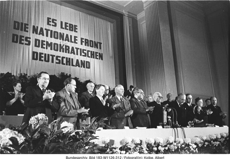 Eine Gruppe von Menschen steht auf der Bühne vor einem großen Transparent mit der Aufschrift "Es lebe die Nationale Front des Demokratischen Deutschland" und applaudiert bei einer offiziellen Veranstaltung.