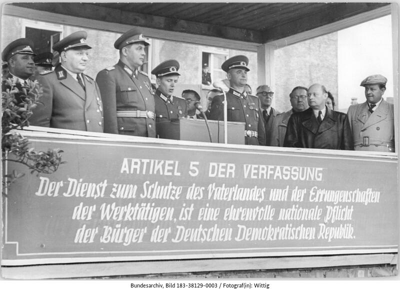 Eine Gruppe von uniformierten und zivilen Beamten steht auf einem Podium hinter einem Schild mit der Aufschrift Artikel 5 der ostdeutschen Verfassung.