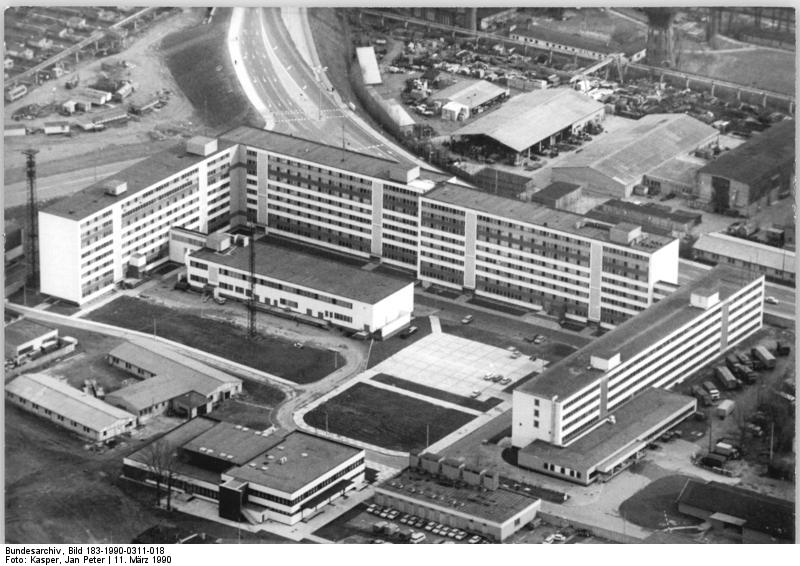 Luftaufnahme eines großen, mehrstöckigen Büro- oder institutionellen Gebäudekomplexes mit angrenzenden Parkplätzen und nahe gelegenen Industrieanlagen.