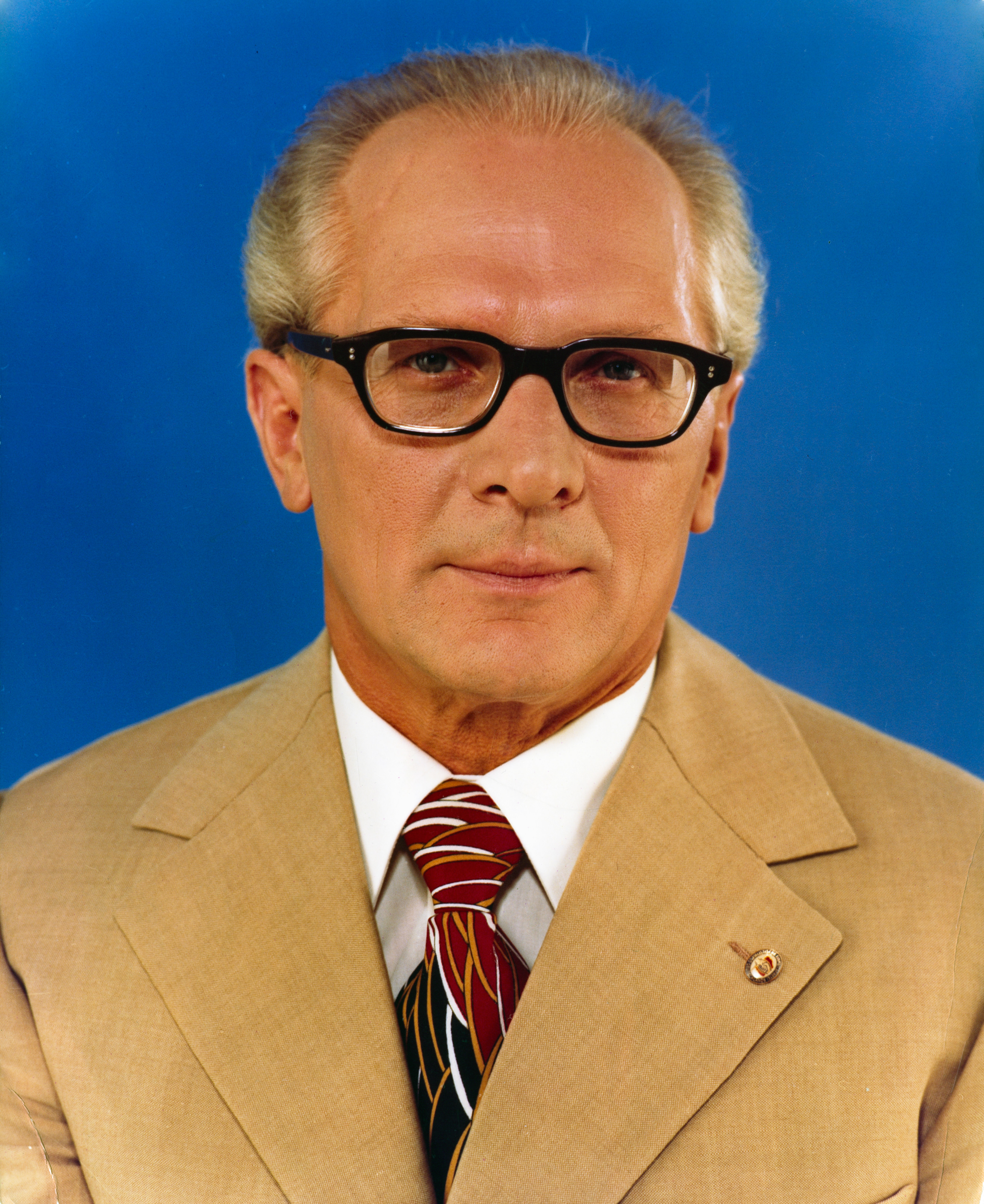 Ein Mann (Erich Honecker) mit kurzen grauen Haaren und Brille trägt einen hellbraunen Anzug, ein weißes Hemd und eine gestreifte Krawatte und posiert vor einem blauen Hintergrund.