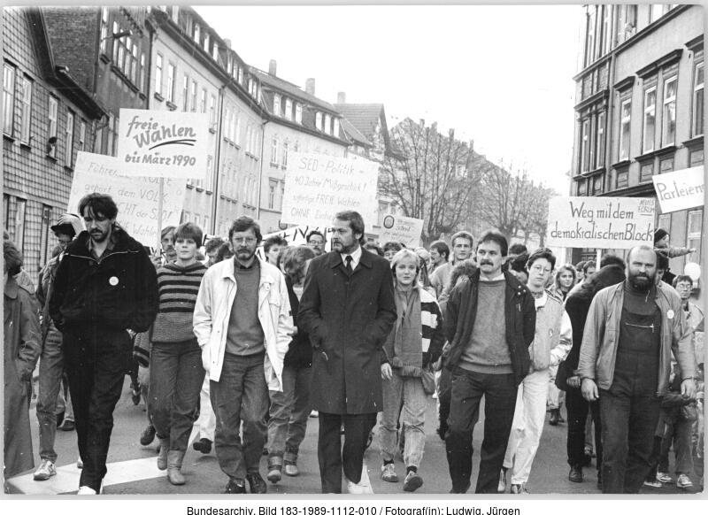 Ein Schwarz-Weiß-Foto einer Gruppe von Menschen, die auf einer Straße in der Stadt marschieren und Protestschilder in deutscher Sprache halten, wahrscheinlich während einer Demonstration in den späten 1980er oder frühen 1990er Jahren.