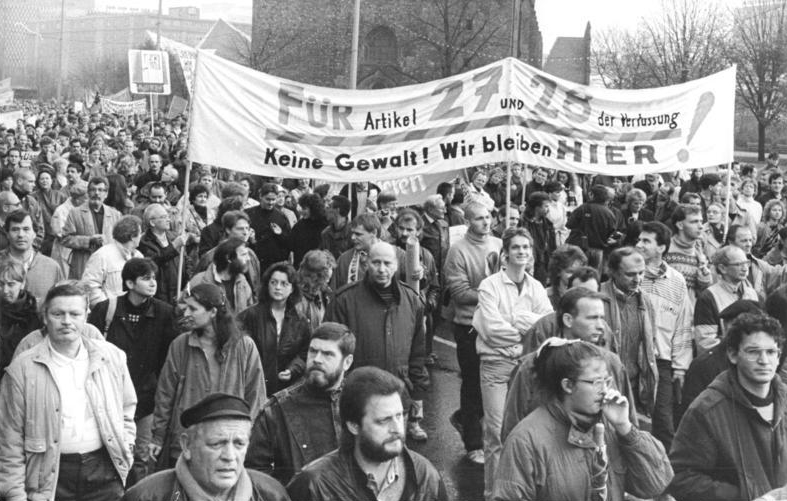 Eine große Gruppe von Menschen demonstriert mit einem Transparent in deutscher Sprache, das zur Gewaltlosigkeit aufruft und auf Artikel 27 verweist, während im Hintergrund Gebäude und Bäume zu sehen sind.