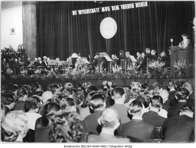 Ein großes Publikum sieht einem Orchester zu, das auf der Bühne spielt, mit einem Dirigenten und einem Redner auf einem Podium; ein Banner und ein Emblem sind auf dem Vorhang hinter ihnen zu sehen.