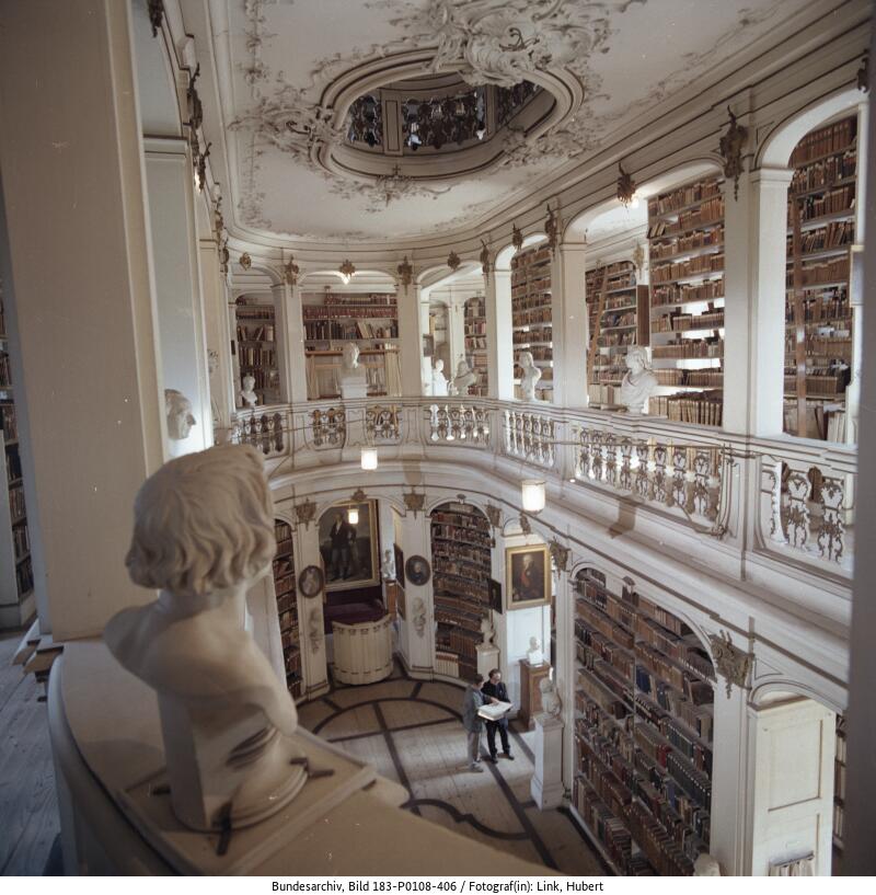Innenansicht einer großen, historischen Bibliothek mit verzierten Balkonen, hohen Decken, Bücherregalen an den Wänden und Marmorbüsten; eine Person liest an einem Tisch darunter.