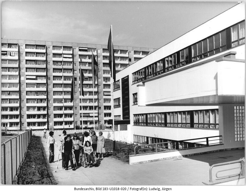 Eine Gruppe von Kindern und Erwachsenen steht vor einem modernen Schulgebäude, mit einem hohen Wohnblock im Hintergrund und Fahnen auf Fahnenmasten in der Nähe.