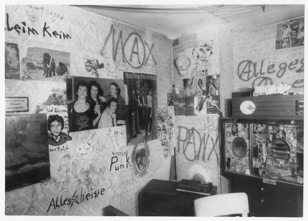 Schwarz-Weiß-Foto eines Zimmers mit mit Graffiti bedeckten Wänden, Bandpostern, Zeichnungen, einem Plattenspieler, Lautsprechern und verschiedenen Gegenständen auf Regalen und Möbeln.