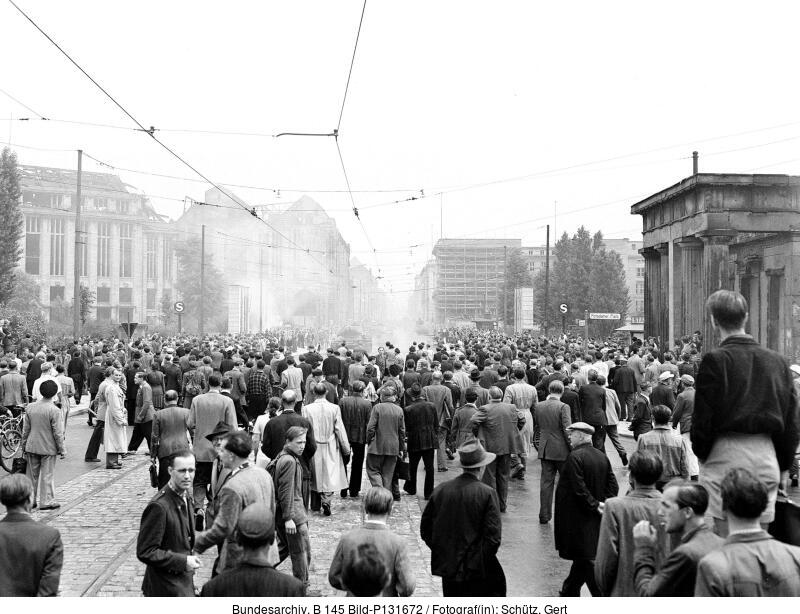Eine große Menschenmenge versammelt sich auf einer städtischen Straße, einige gehen, andere stehen, mit Gebäuden und leichtem Nebel oder Rauch im Hintergrund.