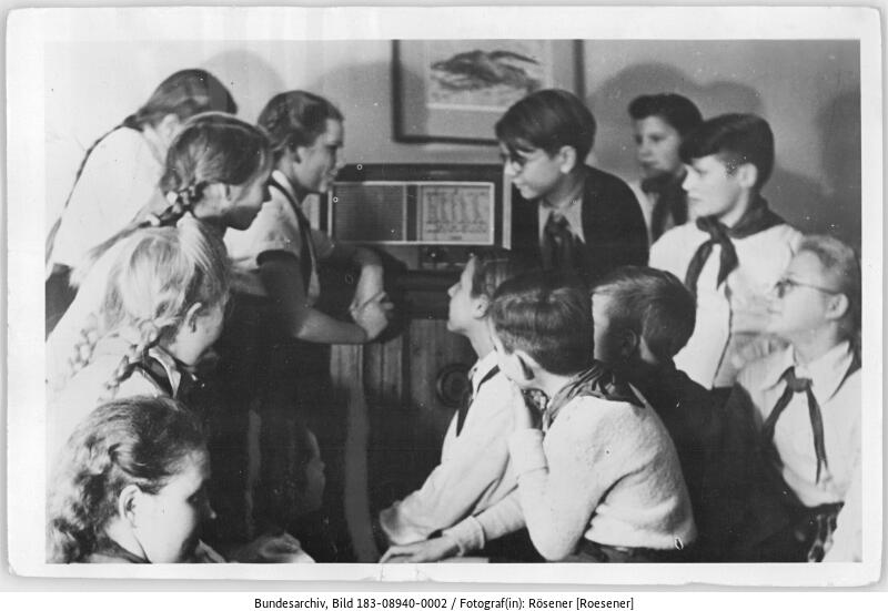Eine Gruppe von Kindern in Uniform versammelt sich um ein Radio und hört in einem Klassenzimmer oder einem Besprechungsraum aufmerksam zu.